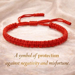 Può includere: Un braccialetto in corda intrecciata rosso brillante con un motivo a nodi macramè e chiusura scorrevole regolabile. Il testo A symbol of protection against negativity and misfortune è stampato sotto il braccialetto in un carattere serif marrone.