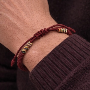 May include: A dark red cord bracelet featuring a sliding knot closure for adjustability. The bracelet includes two decorative accents made of multi-colored thread wrapped around the cord, showcasing shades of yellow, blue, and green.