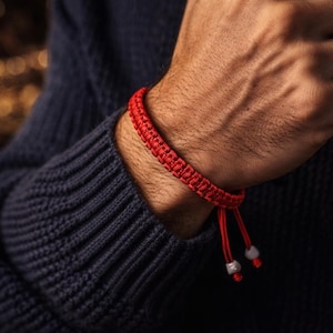 May include: A red woven bracelet with adjustable ties and small silver beads. The bracelet is worn on a person's wrist, contrasting with a dark blue sweater. The bracelet's design is simple and elegant.