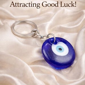 May include: A blue evil eye keychain with a silver keyring and chain. The eye charm has a white and light blue center. The text "Protection & Attracting Good Luck!" is in the background.