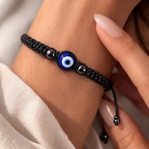 May include: Black woven bracelet featuring a blue and white evil eye charm. The bracelet also includes small, dark gray beads. The adjustable cord allows for a comfortable fit. This bracelet is a stylish accessory.