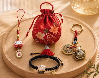 May include: A red drawstring pouch with floral patterns, a red and white ceramic cat charm on a cord, a black braided bracelet with a ceramic cat bead, and a gold-toned keychain featuring a cat figure and a round coin with Chinese characters.