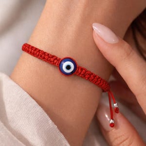May include: Red woven bracelet featuring a blue and white evil eye charm. The bracelet is adjustable with a sliding knot closure and small silver-colored beads. A simple, yet striking piece of jewelry.