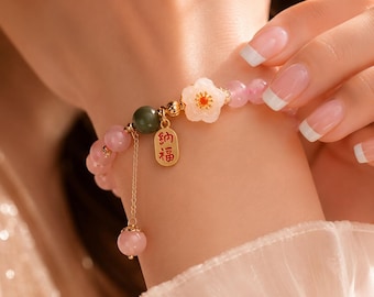 May include: A beaded bracelet featuring translucent pink beads, a single dark green bead, and a white flower-shaped charm with a gold center. A gold rectangular pendant with red Chinese characters hangs from the bracelet alongside a thin gold chain.