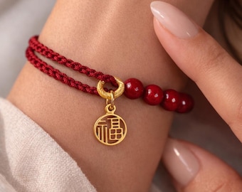 May include: A double-strand red braided cord bracelet featuring a gold-colored circular charm with the Chinese character for good fortune, accented by three smooth, round, deep red beads attached to the cord with a textured gold-colored ring.
