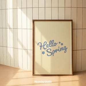 May include: A framed print with a light yellow background and a brown wooden frame. The print features the words "Hello Spring" in a light blue script font, with small flower illustrations. The bottom of the frame has the text "Moonday Studio".