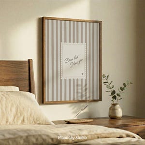 Pode incluir: Uma impressão emoldurada com um fundo listrado e o texto "Dear bed I love you". A moldura é marrom e o fundo tem listras verticais em bege e branco. Uma cama e uma mesa de cabeceira estão em primeiro plano.