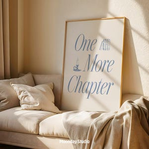 May include: A framed poster with the text "One More Chapter" in blue script, featuring a candle and book illustrations. The poster is displayed on a beige sofa with pillows and a blanket. The frame is gold-colored.