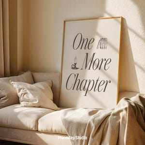 May include: A framed print with the text "One More Chapter" in elegant script, featuring a candle and book illustrations. The artwork is displayed on a beige sofa with pillows and a blanket, creating a cozy reading nook.