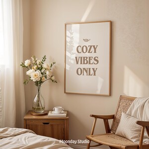 May include: A framed print with the words "COZY VIBES ONLY" in brown lettering hangs on a beige wall. A wooden nightstand holds a vase of white flowers, a cup, and a book. A wooden chair with a woven back is in the foreground.