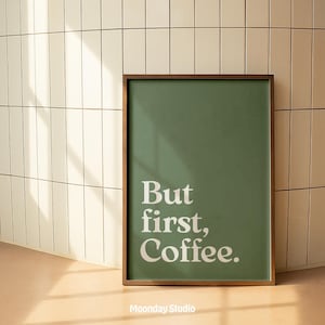 以下が含まれることがあります： 緑色の背景に白い文字で「But first, Coffee」と書かれた額入りのアートプリント。 フレームは濃い茶色です。 プリントは白いタイルの壁に立てかけられています。「Moonday Studio」の文字が下にあります。