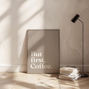 Könnte beinhalten: Ein gerahmter Druck mit dem Text "But first, Coffee." in weißer Schrift. Der Druck lehnt an einer Wand, neben einem Stapel Zeitschriften und einer schwarzen Stehlampe. Der Boden ist aus Holz.