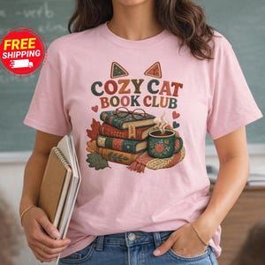 Puede incluir: Una camiseta rosa claro con un gráfico de una pila de libros, una taza humeante y orejas de gato con el texto "COZY CAT BOOK CLUB" en una fuente retro. El diseño incluye hojas de otoño, corazones pequeños y unas gafas sobre los libros.