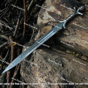 May include: A silver sword with a detailed handle and a patterned blade lies on a weathered wooden surface. The handle has intricate designs, and the blade has a textured pattern. The background includes wooden branches.
