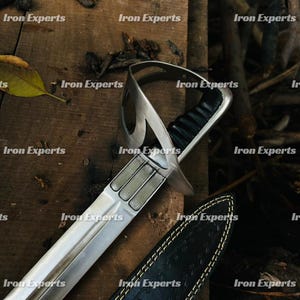 May include: A silver saber with a black handle and a black leather sheath. The blade is shiny and the handle has a curved guard. The sheath is decorated with a stitched pattern. The words "Iron Experts" are visible.