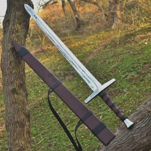 May include: A long, silver sword with a brown leather-wrapped handle and a crossguard. The sword is displayed with its brown leather sheath, which has a black strap. The blade has a textured appearance.