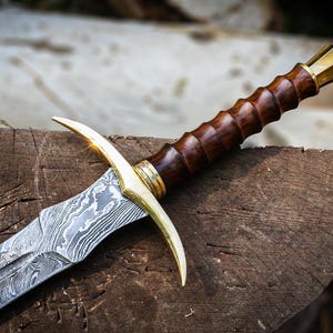 May include: A close-up of a sword with a patterned blade and a wooden handle. The hilt has gold-coloured accents. The sword is displayed on a weathered wooden surface, highlighting its craftsmanship and design.