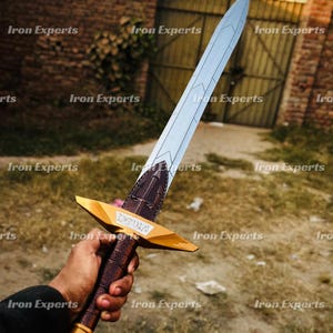 Op de afbeelding: Een decoratief zwaard met een zilveren lemmet en een gouden gevest. Het gevest heeft een bruin, met leer omwikkeld handvat en een gouden pareerstang. Het zwaard wordt in de hand van een persoon gehouden.