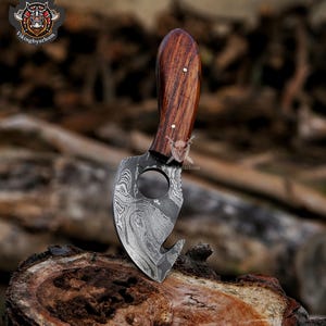 May include: A Damascus steel knife with a wooden handle. The blade has a unique pattern and a curved hook. The handle is made of dark wood and is secured with two rivets. The knife is displayed on a wooden surface.