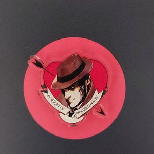 Può includere: Spilla a forma di cuore con un'illustrazione di detective, una freccia e la scritta "A Heart of 99% Gold Alloy". La spilla raffigura un uomo con un cappello e un trench, su un cuore rosso su sfondo scuro.