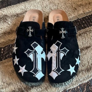 May include: Pair of black clogs featuring a cross and star design. The clogs have a silver cross buckle and a large white cross applique. White star appliques are on the sides. The clogs are size 40.