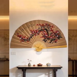 Puede incluir: Gran abanico de pared decorativo con un diseño floral pintado. El abanico presenta un fondo beige con una rama de flores rojas y rosas. Está montado en una pared blanca sobre una mesa de madera oscura.