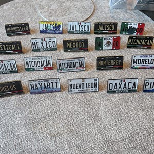 Puede incluir: Una colección de placas de matrícula en miniatura de varios estados mexicanos, incluyendo Jalisco, Michoacán y Oaxaca. Cada placa presenta el nombre del estado y un diseño único, con forma rectangular y acabado brillante. Medidas en centímetros.