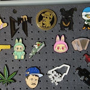 May include: A selection of enamel pins on a grey pegboard. The pins depict diverse designs, including cartoon characters, a gold pistol, a cannabis leaf, and a car. The pins are in various shapes and sizes.