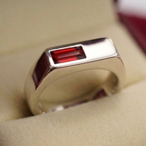 Puede incluir: Un anillo de plata con una piedra preciosa rectangular de color rojo intenso. El anillo está engastado en un bisel rectangular pulido. El anillo se presenta en una caja de joyería roja con un interior color crema. El anillo es de diseño clásico.