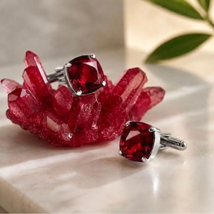 Puede incluir: Un par de gemelos plateados con piedras facetadas de color rojo intenso y forma cuadrada. Un gemelo descansa sobre un grupo de cristales rojos translúcidos y dentados, mientras que el segundo está colocado sobre una superficie lisa y clara.