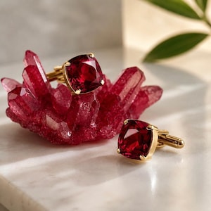May include: A pair of gold cufflinks featuring square-cut, deep red gemstones. One cufflink rests atop a cluster of jagged, translucent red crystals, while the other sits beside it, showcasing the polished gold metal backing and prong settings.