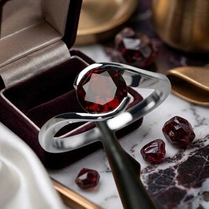 May include: A silver ring featuring a large, faceted red gemstone. The ring is presented with loose red gemstones and a maroon velvet ring box. The ring's design has a flowing, modern style.