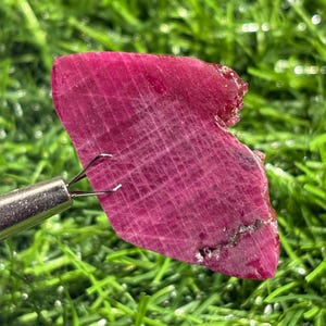 Puede incluir: Una pieza plana e irregular de rubí en bruto de color rojo magenta intenso. La superficie muestra un patrón visible en forma de cuadrícula de líneas entrecruzadas y una textura cristalina rugosa con una pequeña inclusión oscura en el borde.