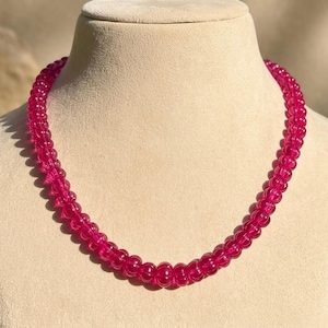 May include: A vibrant pink beaded necklace displayed on a mannequin. The necklace is composed of numerous small, round beads, creating a continuous, eye-catching strand. The beads have a glossy finish, reflecting light and adding to the necklace's appeal. The necklace is a statement piece.
