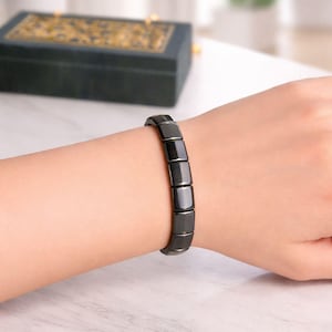 Op de afbeelding: Een zwarte armband met rechthoekige schakels wordt om een pols gedragen. De armband is gemaakt van een glanzend, donker materiaal. De achtergrond bevat een decoratieve doos en een plant.