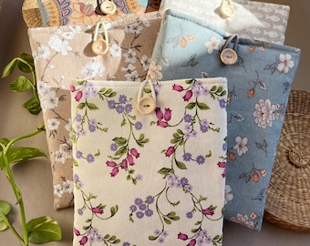 Pode incluir: Uma coleção de capas de livros de tecido com estampas florais. Cada capa possui um fecho de botão de madeira e é feita de tecidos de cores diferentes, incluindo bege, azul e branco. Uma cesta tecida e vegetação também estão presentes.