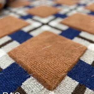 Puede incluir: Primer plano de una alfombra con un patrón geométrico. El diseño presenta grandes formas de diamante marrón sobre un fondo de cuadrados azules, blancos y marrones. Las letras "RAC" son visibles en la esquina inferior izquierda.
