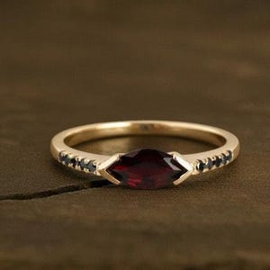 以下が含まれることがあります： 中央にマーキス型の濃い赤色の宝石、両側に小さな黒い石をあしらったゴールドリング。リングは、そのエレガントなデザインと職人技を示す、濃い木製の表面に置かれています。