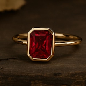 Op de afbeelding: Een gouden ring met een grote, rechthoekige, dieprode edelsteen. De edelsteen is gezet in een gouden bezel en de ringband is een eenvoudig, gepolijst goud. De ring wordt gepresenteerd op een donkere, getextureerde houten ondergrond.