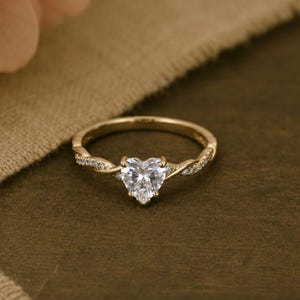 May include: A gold ring featuring a central heart-shaped clear gemstone. The band has a twisted design on both sides of the center stone, with small clear accent stones embedded along the twisted sections of the metal band.