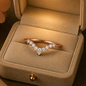 May include: A rose gold V-shaped ring featuring a series of small round clear stones and a single pear-shaped clear stone at the center point. The ring is displayed inside a beige velvet-lined jewelry box with an integrated LED light.