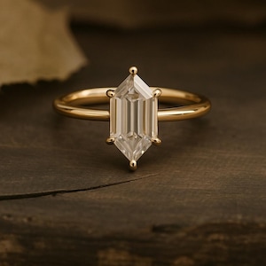 Puede incluir: Un anillo de oro con una gema clara de corte hexagonal. El anillo presenta una banda fina y pulida y seis pequeñas garras que aseguran la gema. El anillo está sobre una superficie de madera oscura.