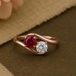 Könnte beinhalten: Ein Ring aus Roségold im Bypass-Design mit einem runden roten Rubin und einem runden weißen Diamanten. Die beiden Steine sind nebeneinander in einer zarten, geschwungenen Ringschiene gefasst, die sich elegant um jeden Edelstein legt.