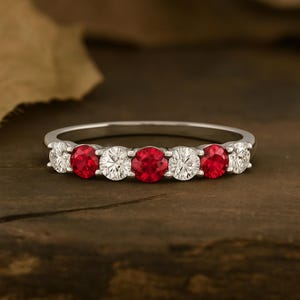 以下が含まれることがあります： 赤と透明な宝石が交互に並んだシルバーリング。 リングには、5つの赤い宝石と4つの透明な宝石が、シルバーのバンドにセットされています。 宝石は丸く、均等に配置されています。