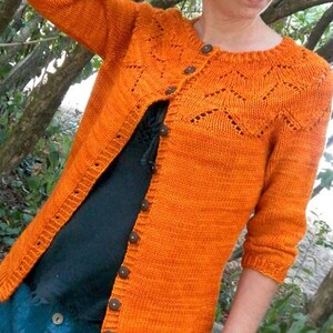 May include: An orange knitted cardigan with a round neckline and button closure. The cardigan features a decorative knit pattern on the yoke and is worn over a black top and blue pants. The sleeves are three-quarter length.