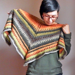 May include: A hand-knitted shawl in a triangular shape, with a striped pattern in orange, green, grey, and white. The shawl has an openwork design and is held up to display its colours and texture.