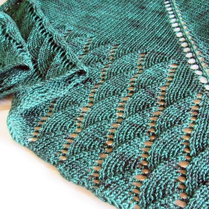 May include: A close-up of a hand-knitted shawl in shades of teal and dark green. The shawl features a detailed openwork pattern with a scalloped edge and a delicate, lacy design. The texture of the yarn is visible.