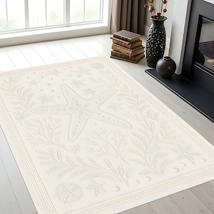 Puede incluir: Alfombra rectangular de color crema con diseño marino. La alfombra presenta una gran estrella de mar en el centro, rodeada de algas en relieve, estrellas de mar más pequeñas y otros elementos marinos. Un borde decorativo enmarca el diseño.
