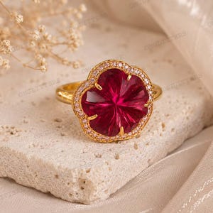 May include: A gold ring with a large, ruby-red, flower-shaped gemstone, encircled by smaller pink stones. The ring is displayed on a textured, off-white surface, against a soft, neutral-toned backdrop. The ring's design is intricate and eye-catching.