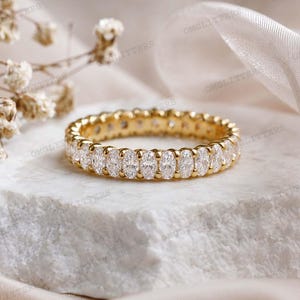 Può includere: Un anello eternity dorato con una fila continua di pietre preziose ovali e trasparenti. L'anello è appoggiato su una superficie di pietra bianca, con morbidi accenti floreali sfocati e tessuto bianco sullo sfondo.